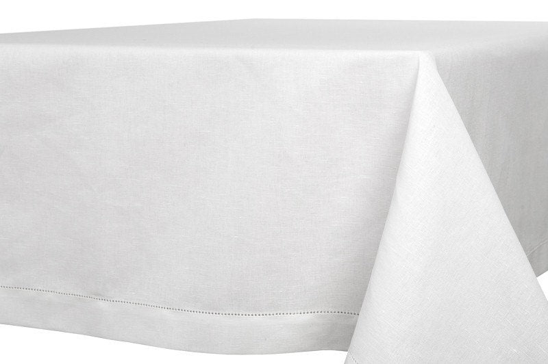 White linen tablecloth draped over a rectangular table, showing its hemstitched border and wide hem.