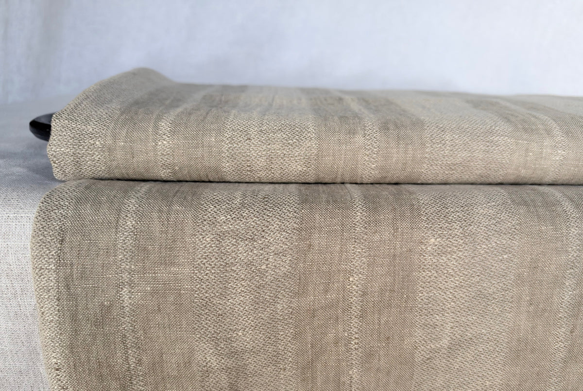 A folded piece of textured linen fabric in a warm taupe-beige color, displayed on a matching surface against a neutral light gray background.