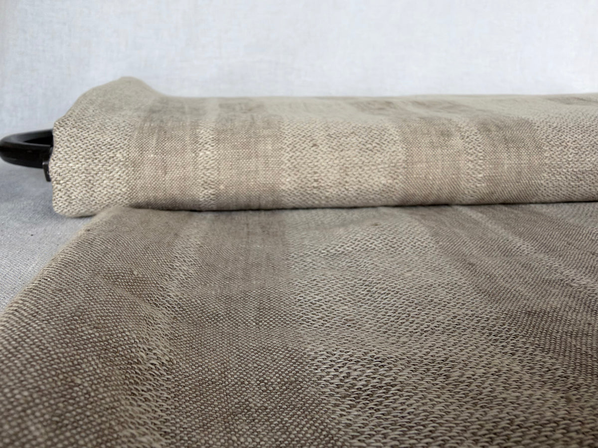 A close-up of a rolled woven fabric or rug in a taupe-beige color, showing its textured, basketweave-like pattern against a neutral gray background.