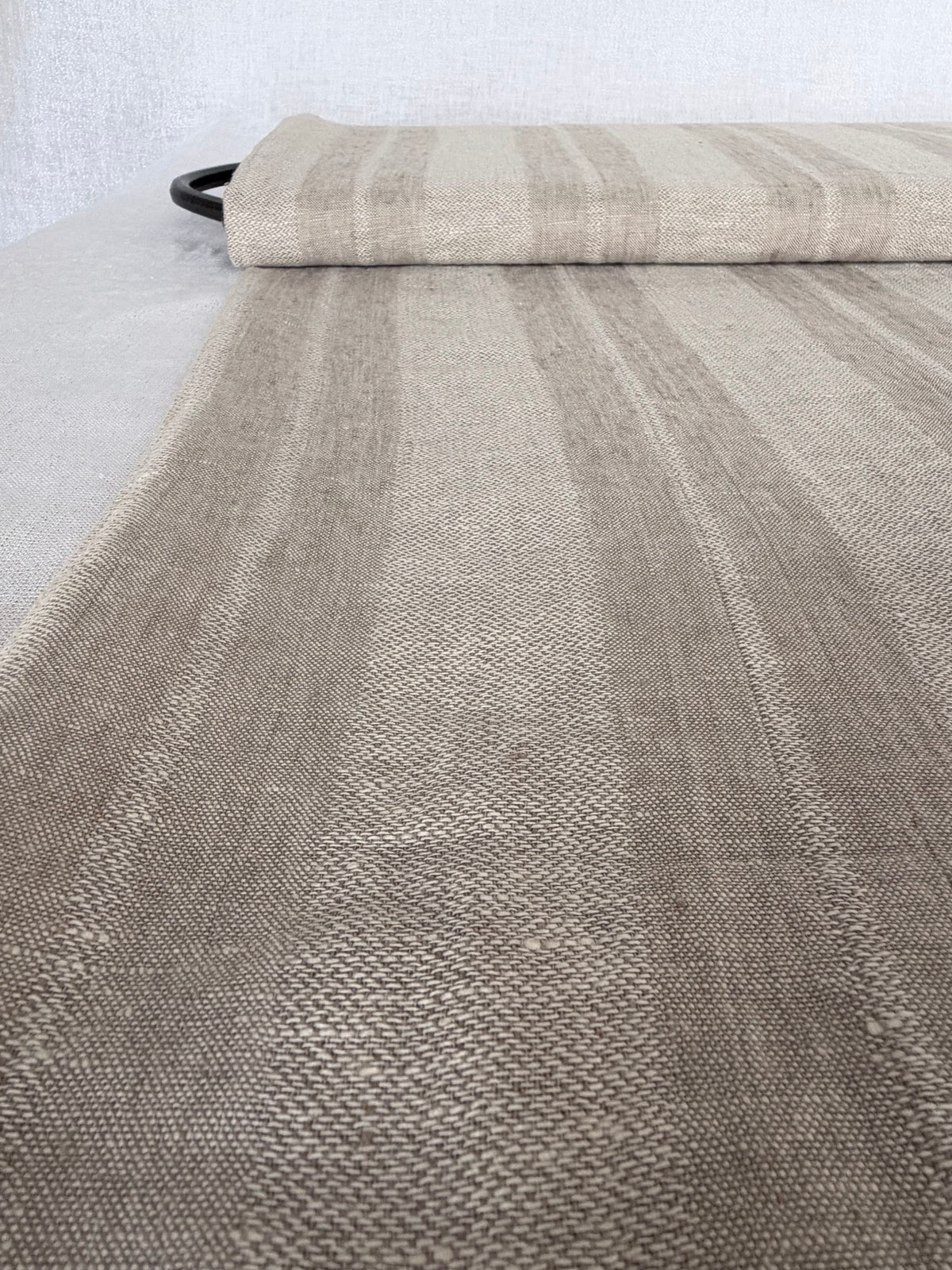 A close-up of a woven rug or textile laid out on a light surface, with a rolled section at the top. The color identity is a neutral mix of taupe, beige, and light brown in a subtle striped pattern.