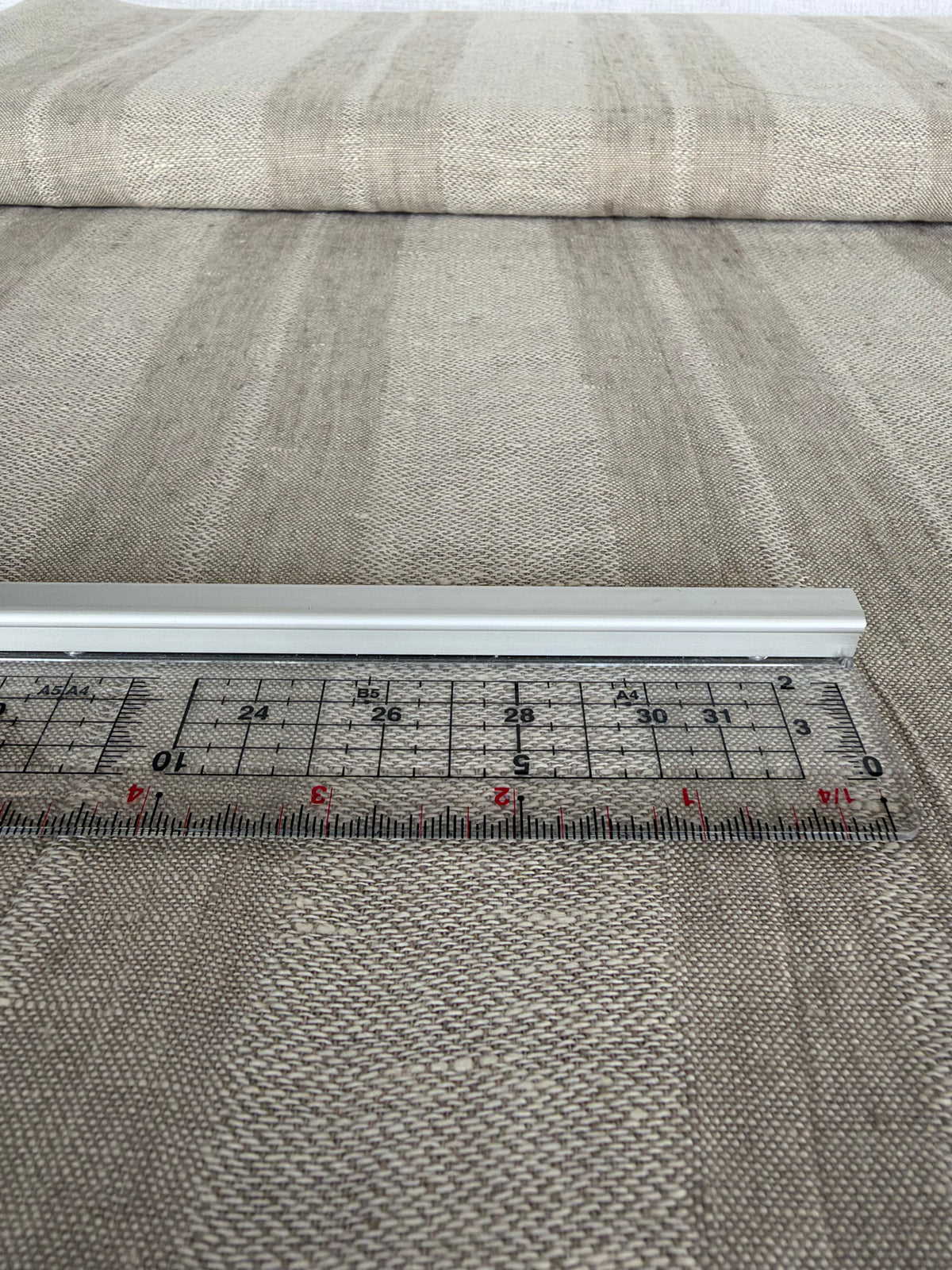 A close-up of a woven fabric with vertical stripes in beige, gray, and brown tones, with a metal ruler placed horizontally to show scale.