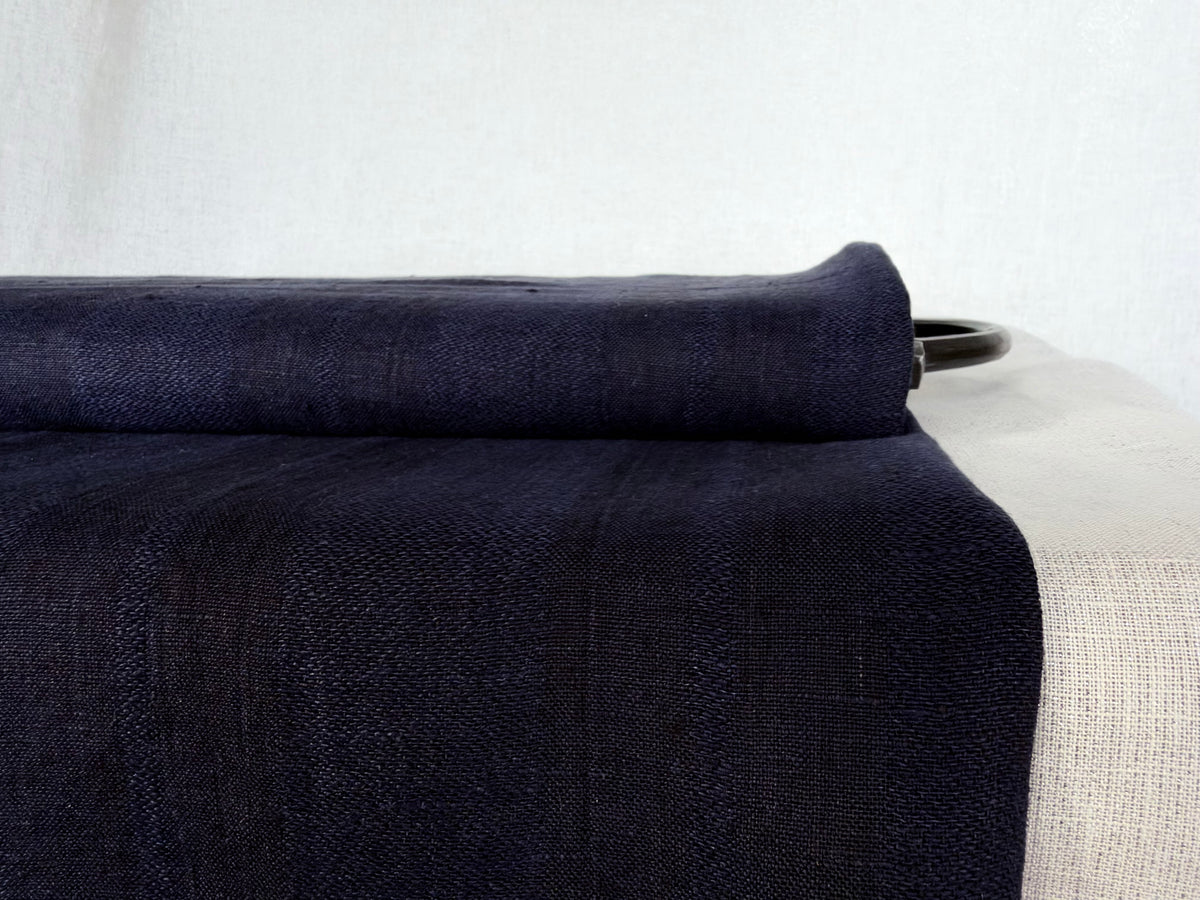 A close-up of a folded dark navy blue woven fabric, likely a linen throw blanket or textile, draped over a light-colored upholstered chair with a black metal ring visible behind it.