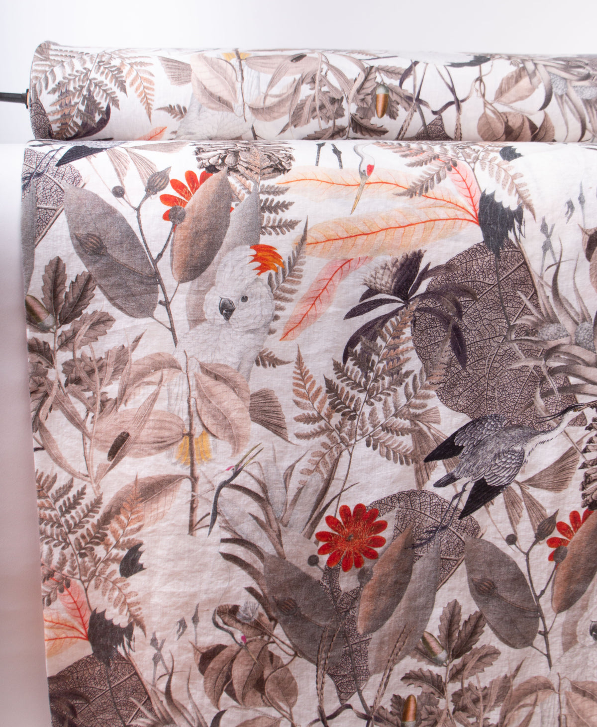 A close-up of a rolled fabric or textile with an all-over botanical and bird print. The pattern features muted gray and brown leaves, ferns, and birds with bright red-orange flower accents on a light off-white background.