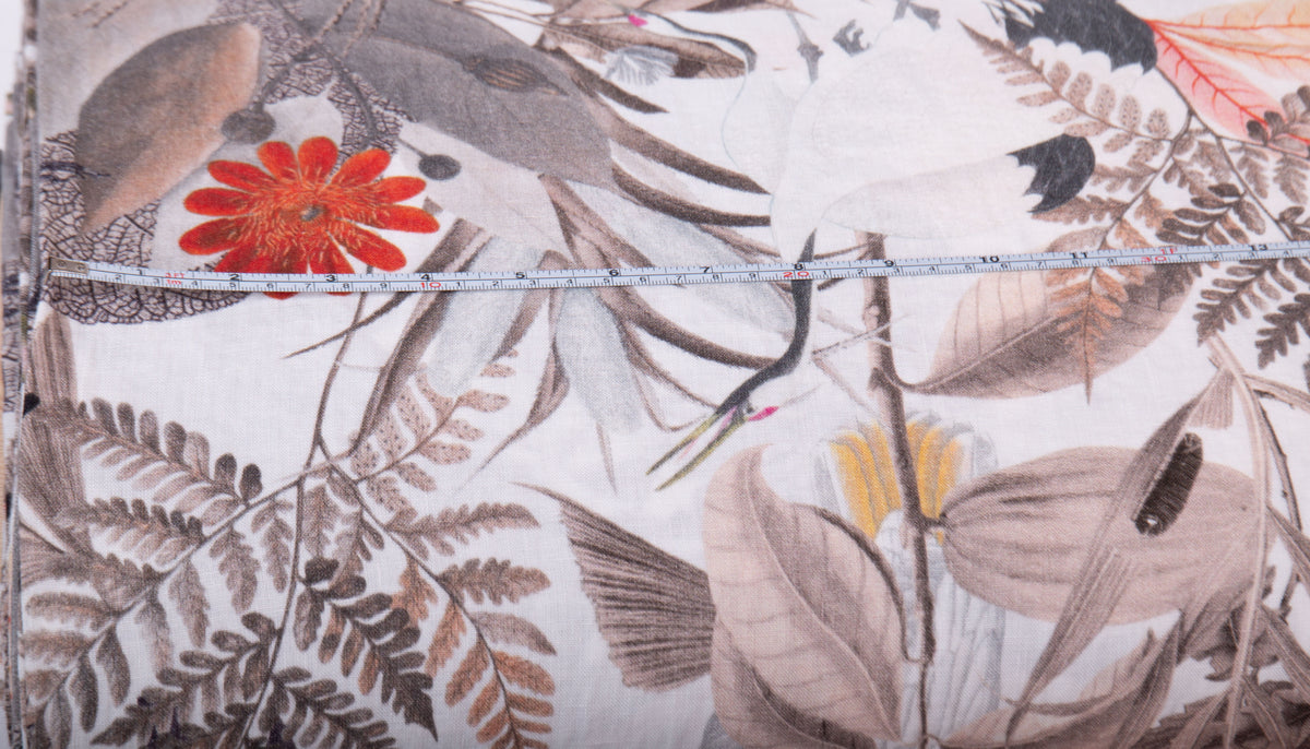 A close-up of a fabric or wallpaper pattern featuring botanical leaves, birds, and a bright red flower on a light background, with a measuring tape laid horizontally across the center.