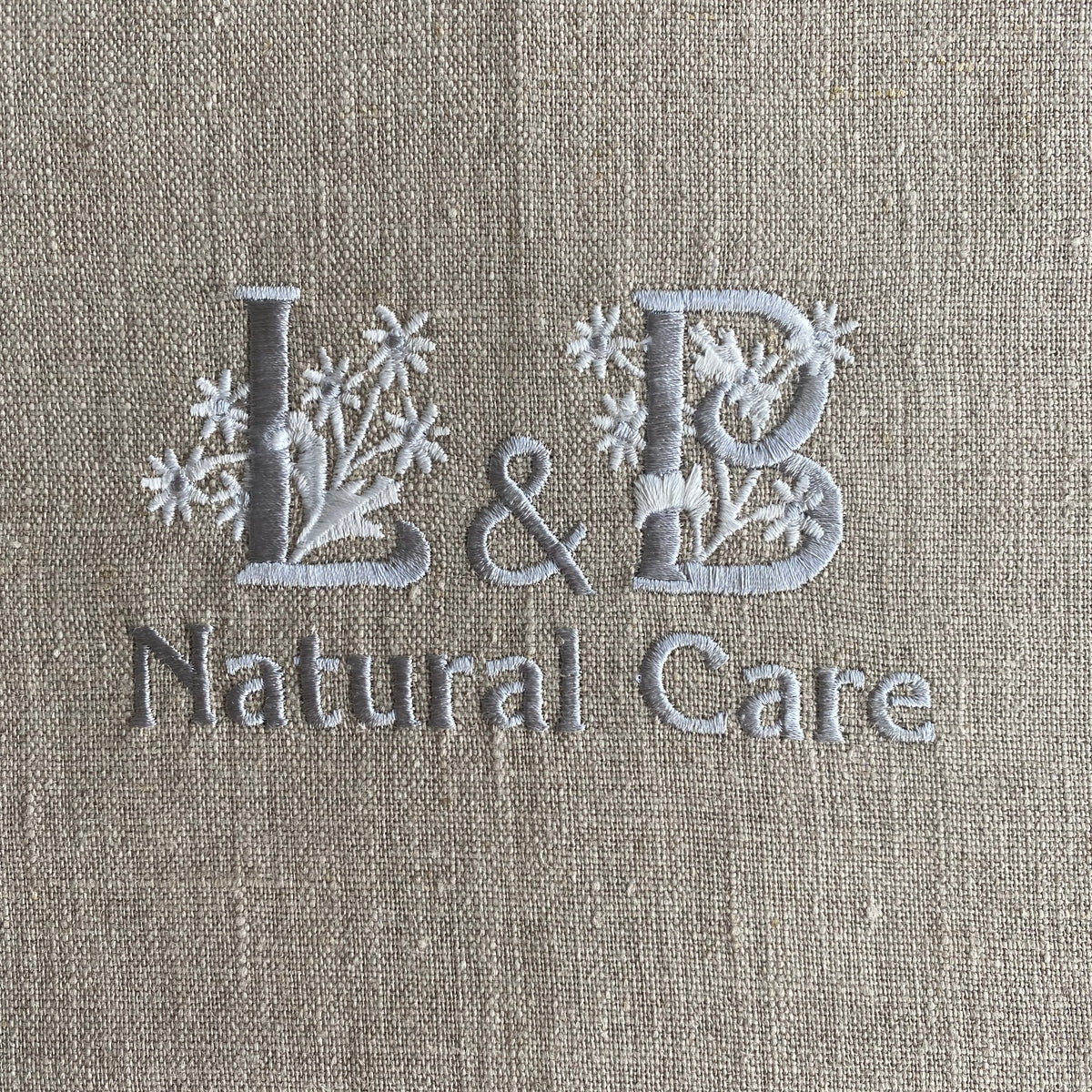 Close-up of a beige linen-textured surface with embroidered gray lettering reading 'L & B Natural Care' and small floral motifs around the letters.