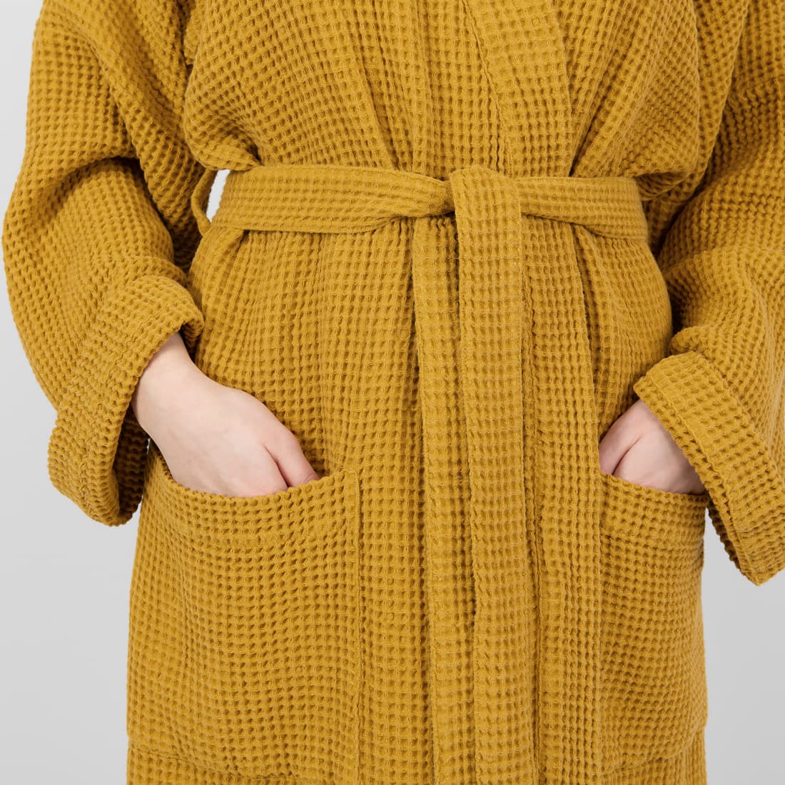 A close-up of a mustard yellow waffle-knit bathrobe with a tie belt and front pockets, worn by a model whose hands are tucked into the pockets.