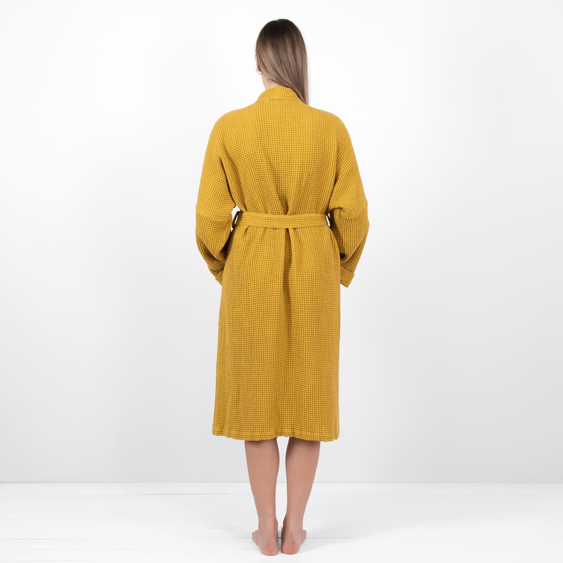 A woman viewed from behind wearing a mustard yellow waffle-knit bathrobe with a matching tie belt, standing against a plain white studio background.