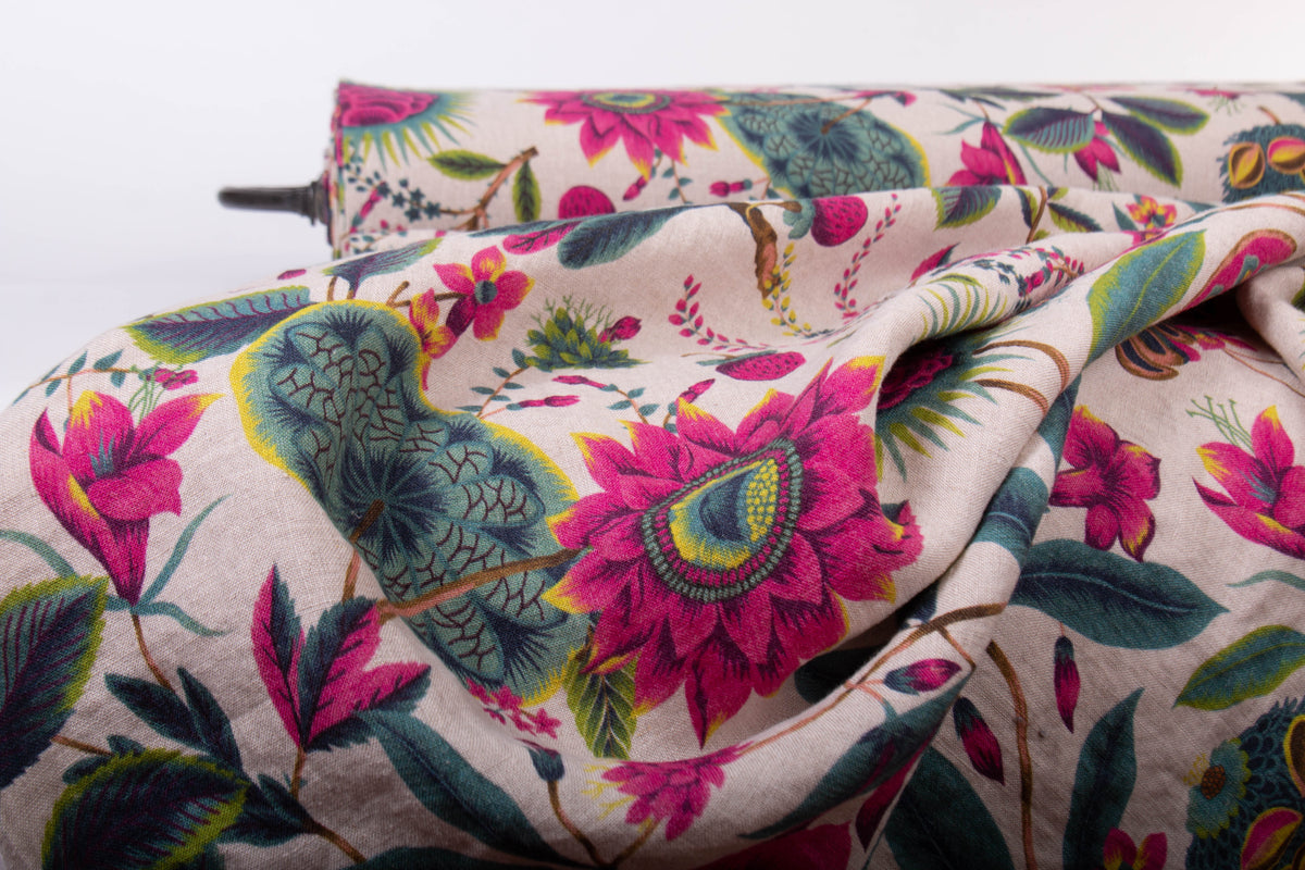 A close-up of draped fabric with a dense floral and botanical print on a light beige background, featuring bright pink flowers, teal and green leaves, and small yellow accents.
