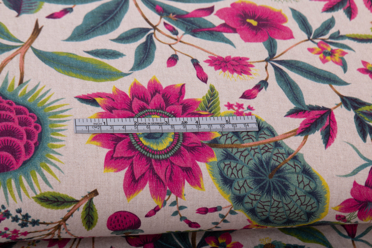 A close-up of a floral fabric with a large-scale botanical print featuring bright pink flowers, teal and green leaves, and yellow accents on a light beige background, with a silver metal ruler placed horizontally for scale.