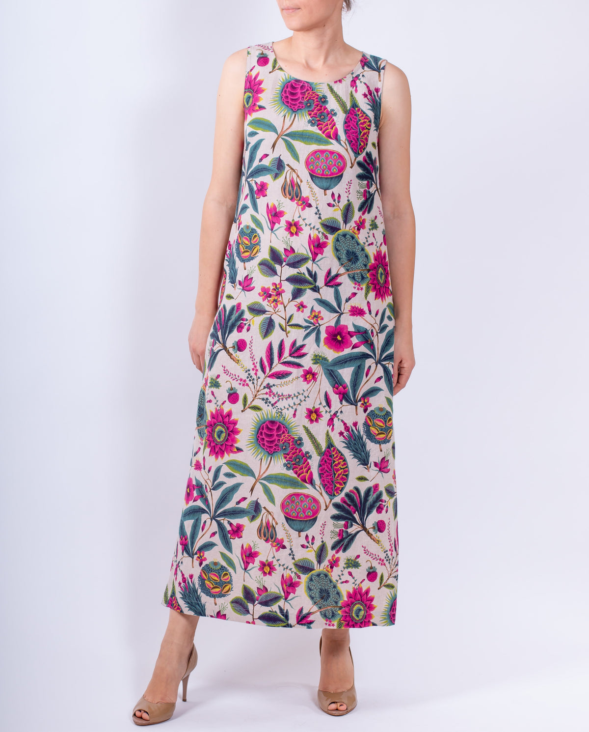 A woman modeling a sleeveless maxi dress with a vibrant, multicolored botanical and floral pattern on a light cream background, set against a plain light gray studio backdrop.