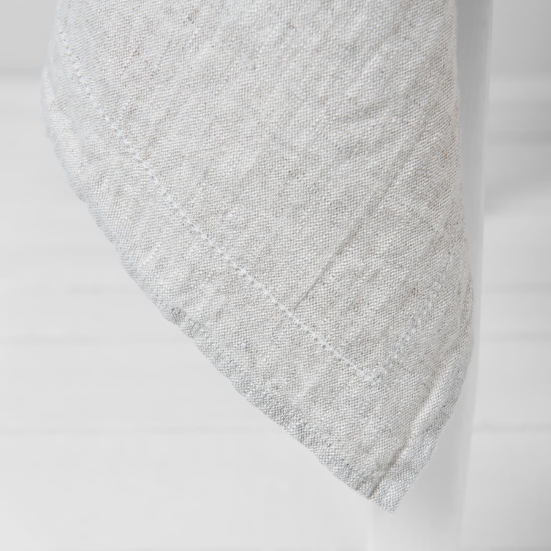 A close-up of a light gray textured fabric, likely a linen tablecloth or napkin, draped over a white stand against a white background.