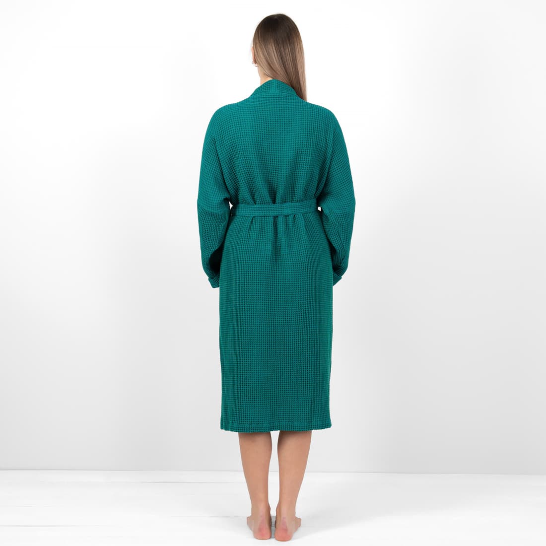 A woman stands with her back to the camera wearing a long-sleeved, belted green robe with a textured waffle pattern, set against a plain white studio background.
