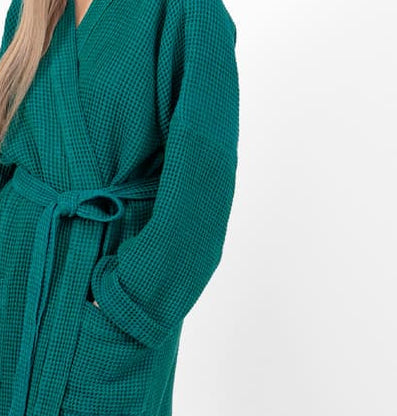 A woman modeling a long, dark teal green waffle-knit bathrobe with a matching tie belt, standing against a plain white studio background.