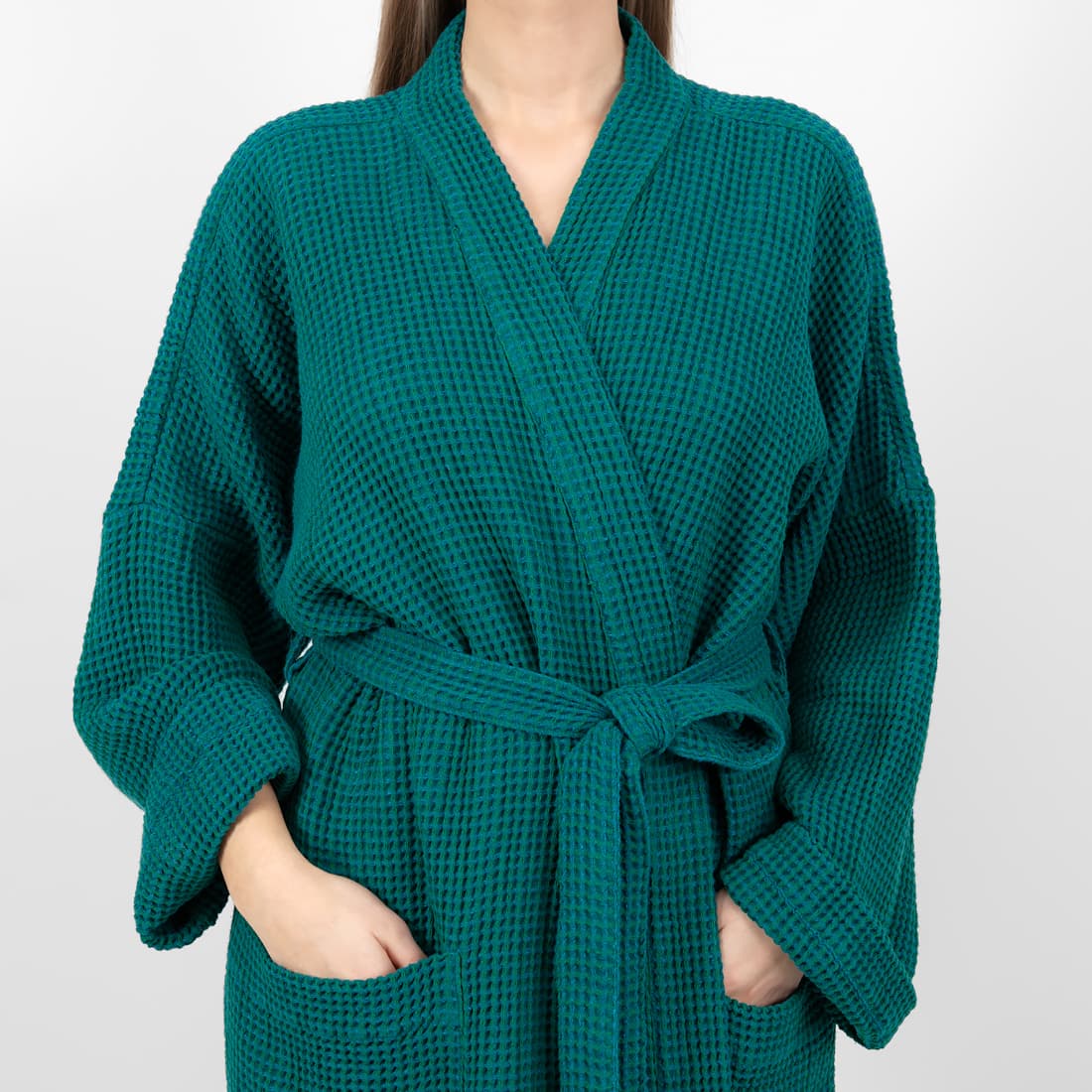 A woman wearing a dark teal green waffle-knit bathrobe with a matching tie belt, shown from the neck down against a light gray background.