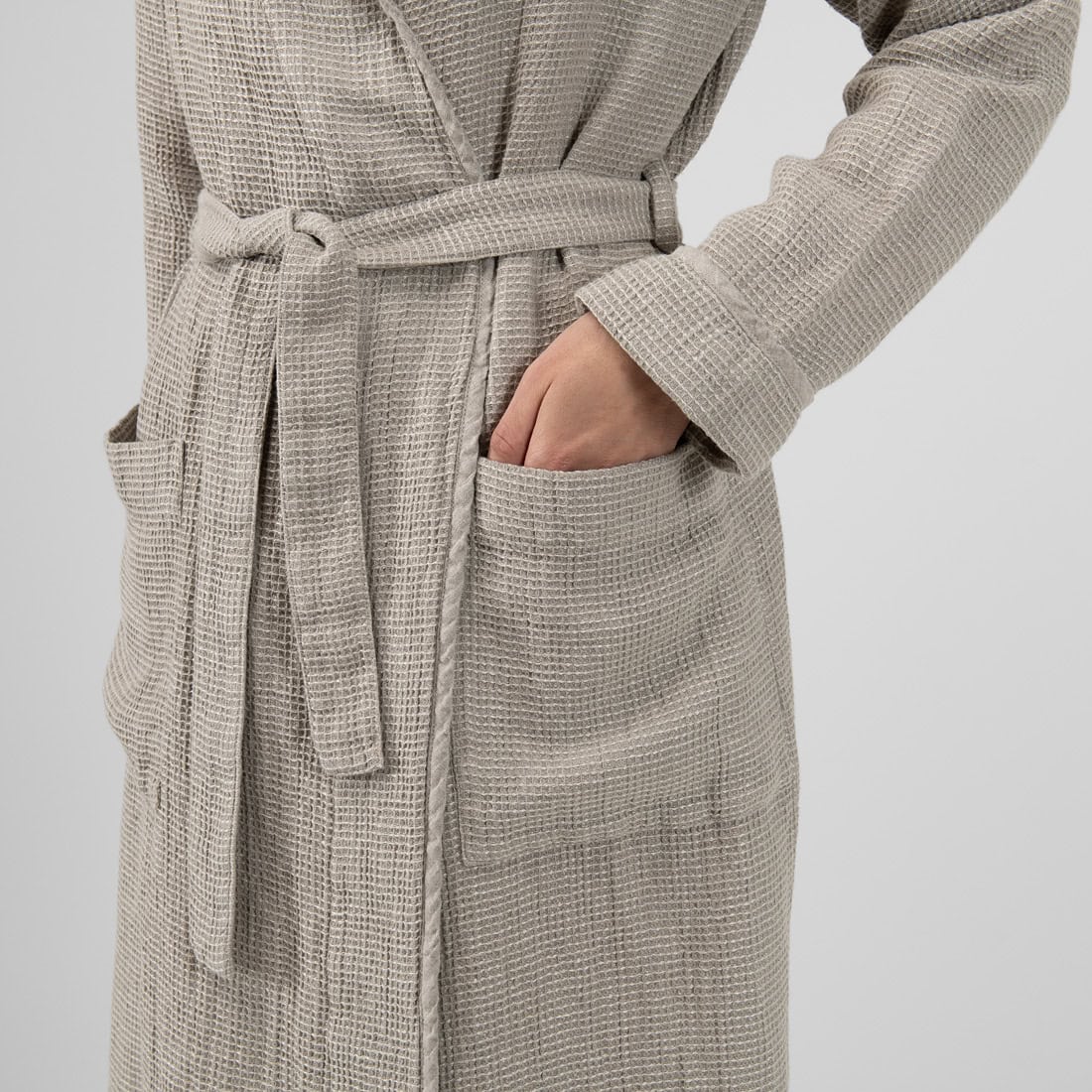 A close-up of a person wearing a textured waffle-knit bathrobe in a light taupe-beige color, with one hand tucked into a front pocket against a plain light gray background.