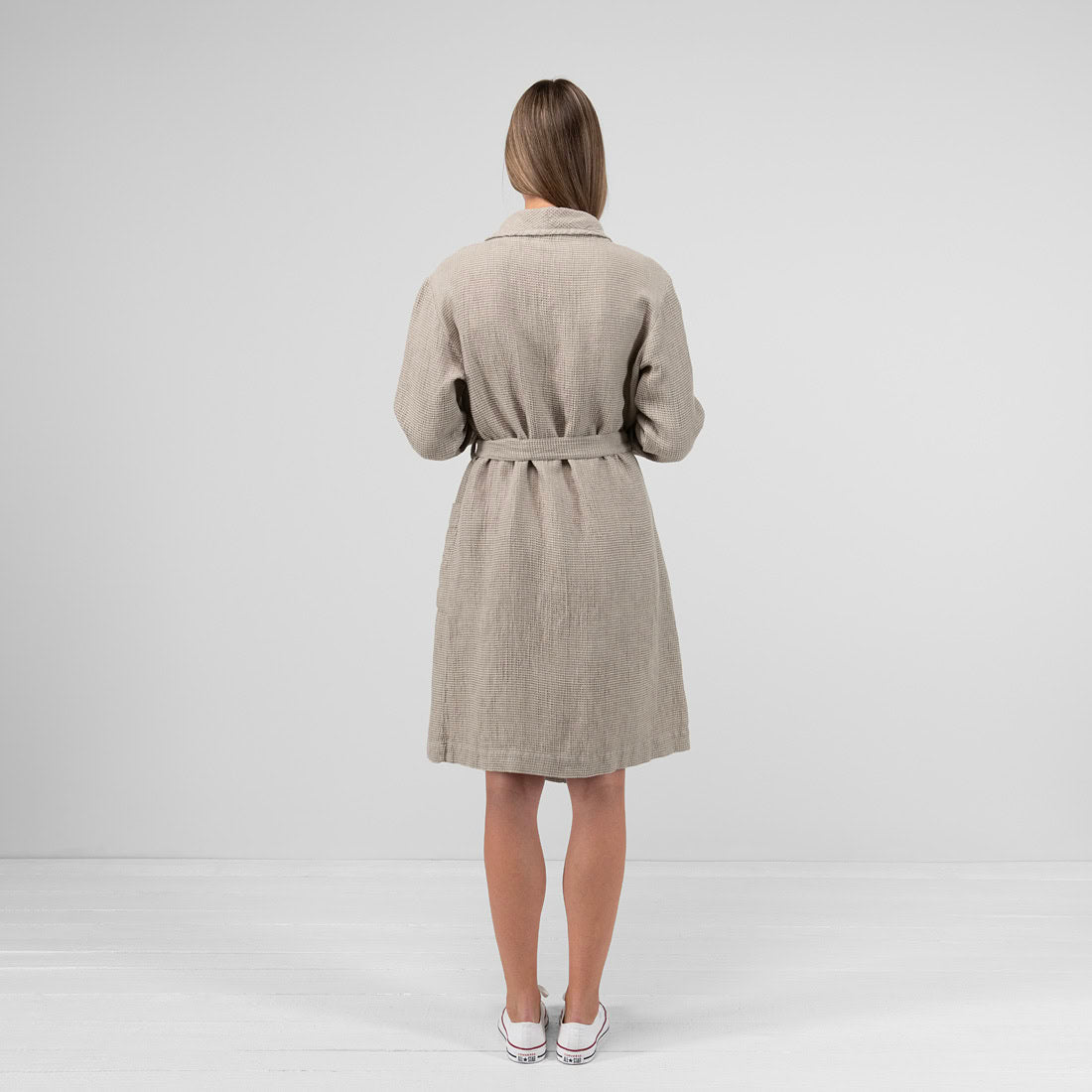 A woman viewed from behind wearing a light beige linen belted coat or kimono-style jacket, standing against a plain light gray studio background.