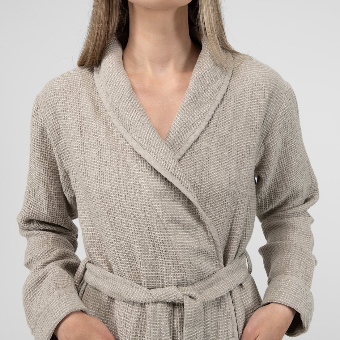 A woman wearing a beige waffle-knit bathrobe with a shawl collar and matching tie belt, shown from the neck down against a light gray background.