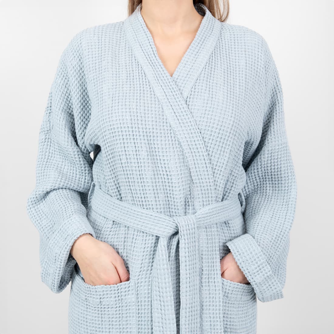A woman wearing a light blue waffle-knit bathrobe with a matching tie belt and front pockets, shown from the neck down against a plain light gray background.