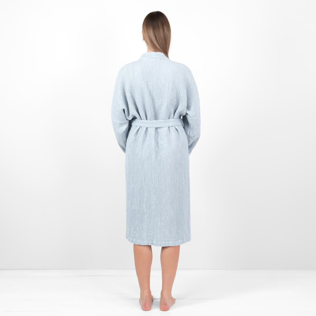 A woman viewed from behind wearing a light blue and white checkered waffle-textured bathrobe, standing against a plain white studio background.